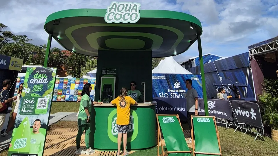 Estande da AQUACOCO na WSL Challenger Series em Maresias, com cobertura verde em ondas, balcão e espreguiçadeiras, e visitantes no atendimento.