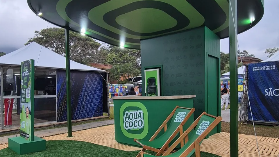 Estande da AQUACOCO visto de lado, com parede estampada com a marca e totem da promoção Deu Onda.